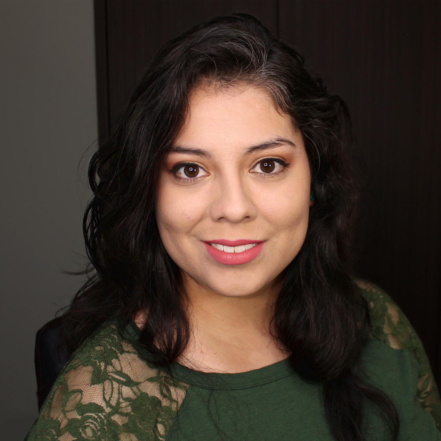 A brown-skinned woman with long wavy dark brown hair is looking straight at the camera and smiling..